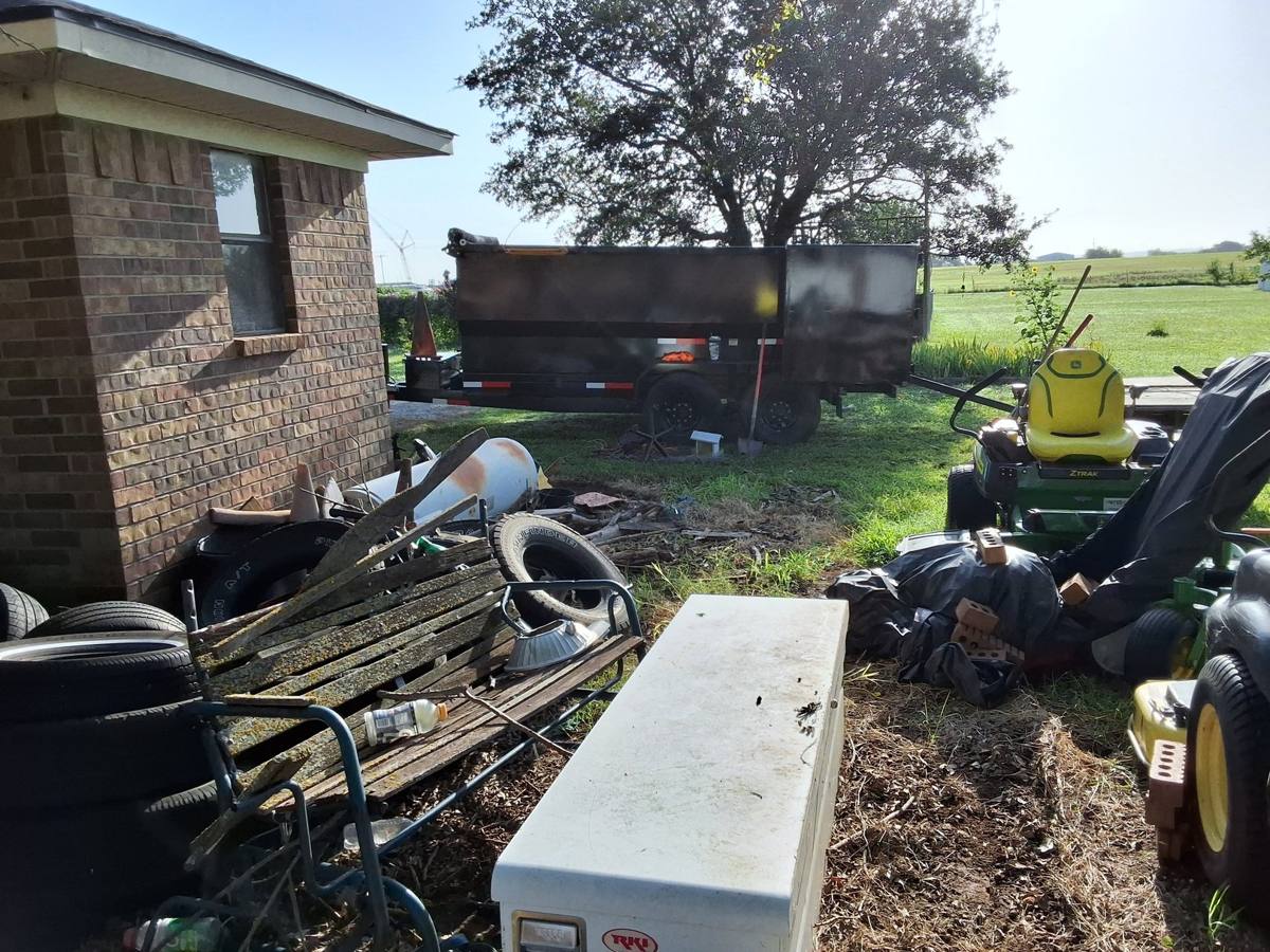Dump trailer parked beside a brick building, with tires, furniture, and equipment gathered nearby